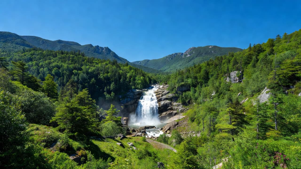 A cascading waterfall flows down a rocky cliffside surrounded by a dense, vibrant green forest. The vast mountain range rises against a bright blue sky under clear and sunny weather conditions.