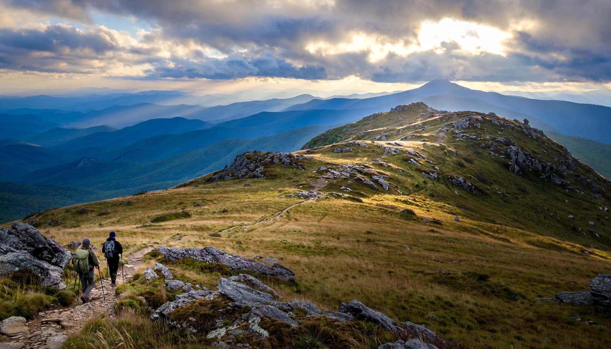 Two hikers follow a rocky trail across a grassy ridge toward distant blue mountains.