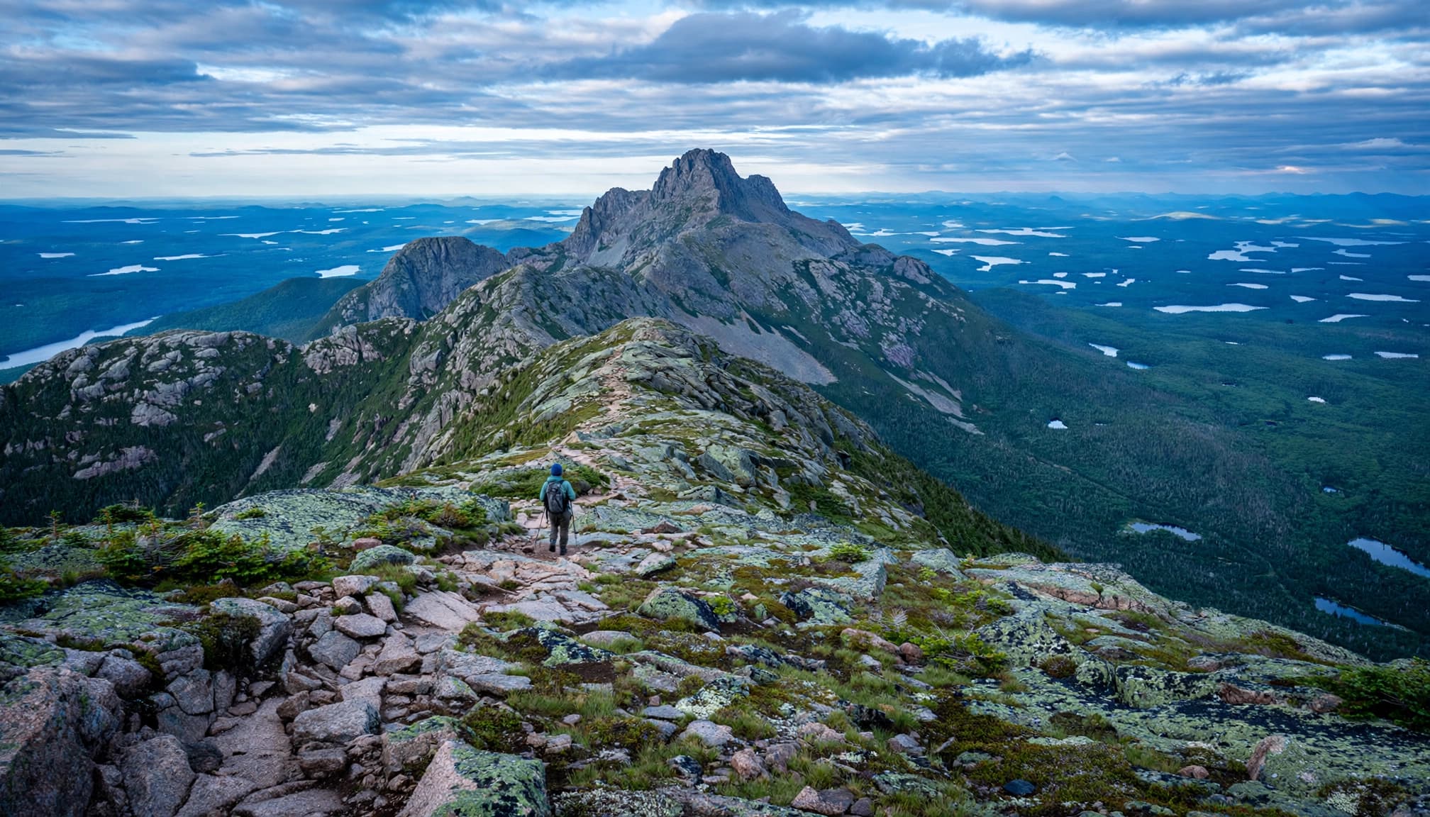 A lone hiker crosses rocky alpine terrain toward a summit above distant lakes.