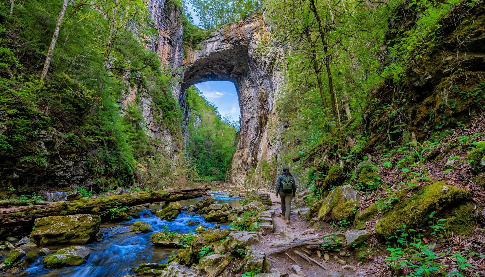 A hiker walks toward a huge limestone arch on a forest trail.