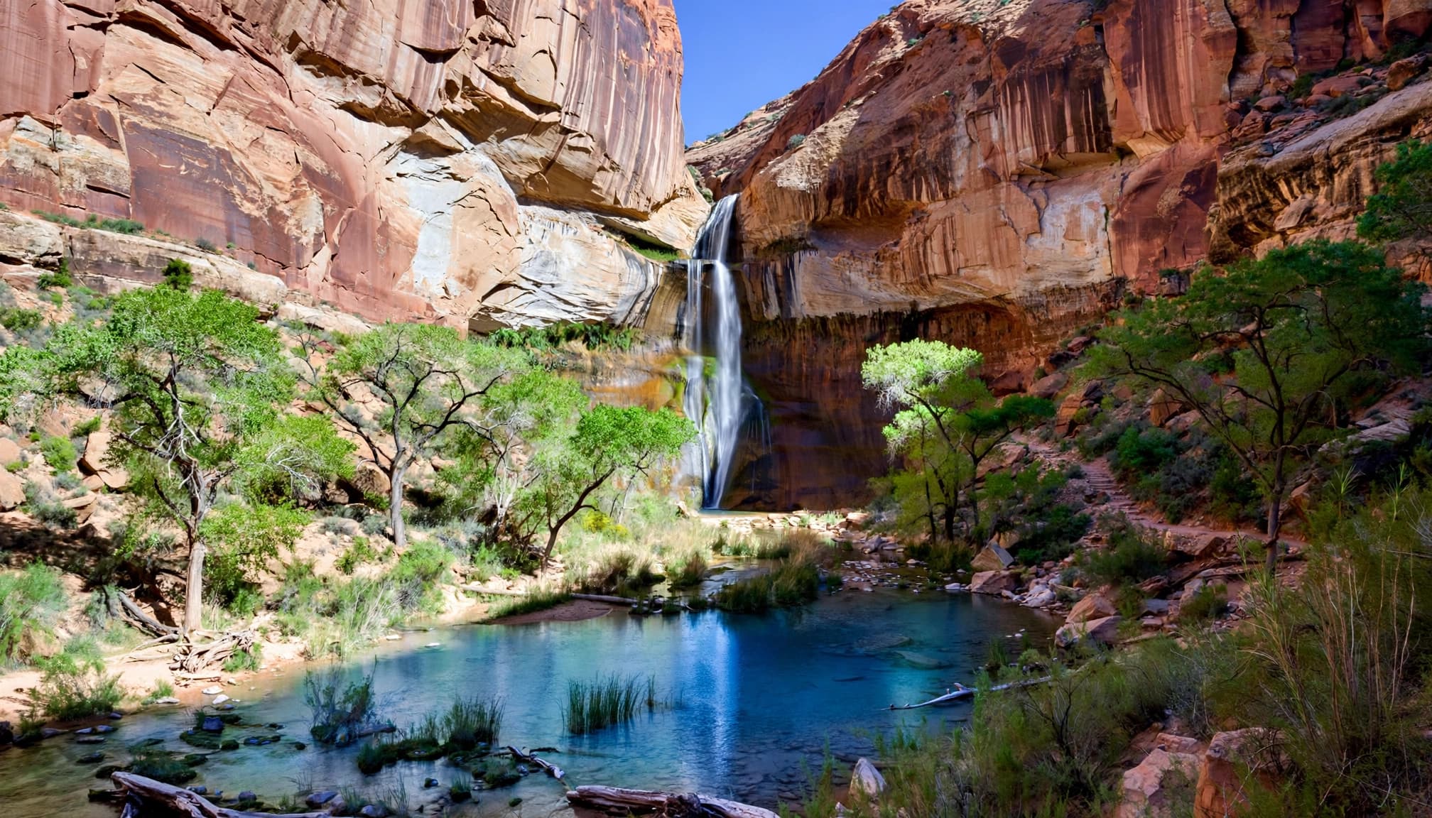 A tall waterfall cascading into a blue pool surrounded by red sandstone cliffs and green trees.