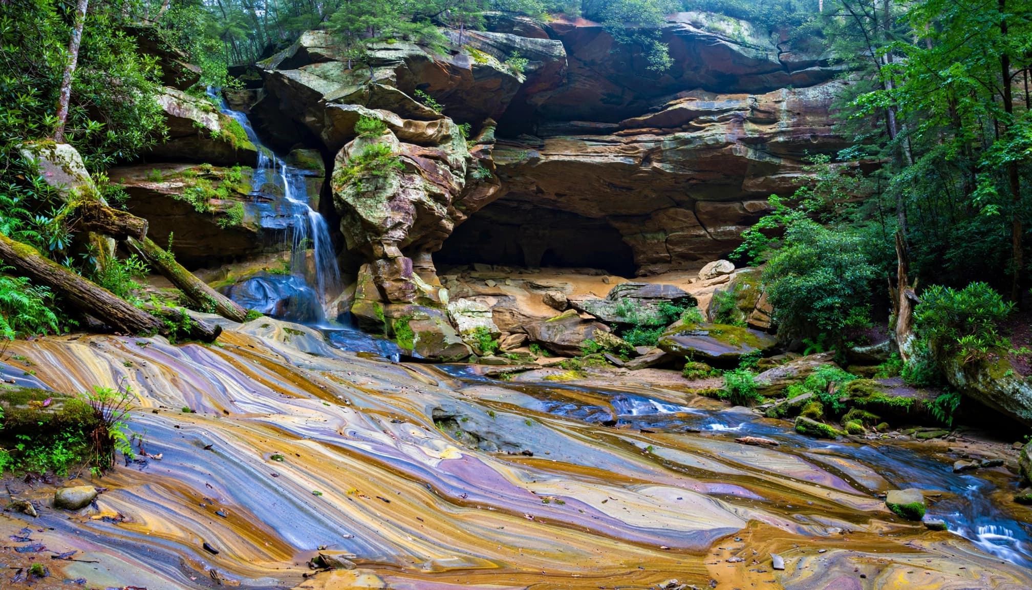 A scenic waterfall cascading over layered sandstone rock and multicolored sand surrounded by a lush forest.